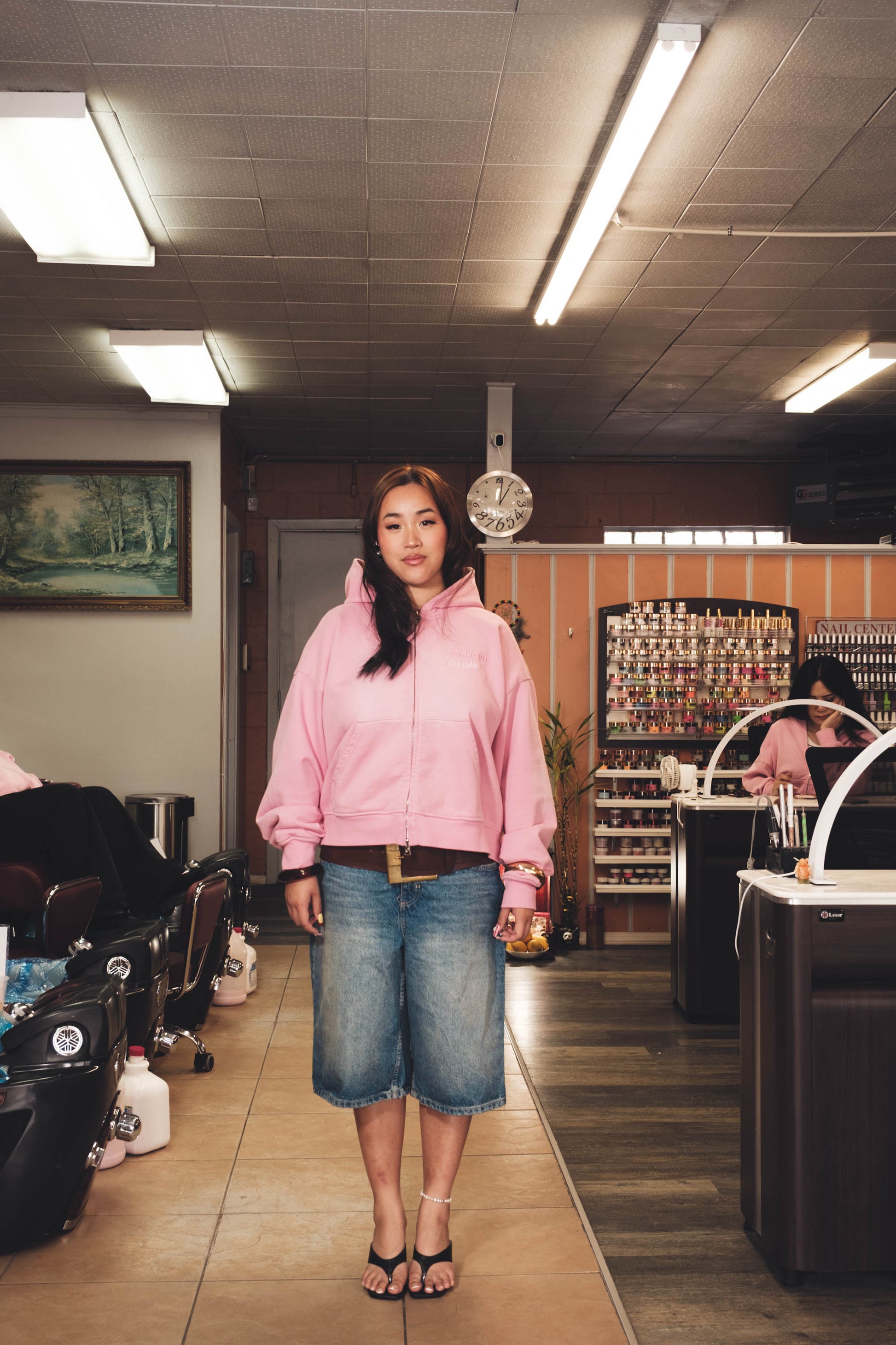 Woman in a pink hoodie and denim shorts standing in a salon.